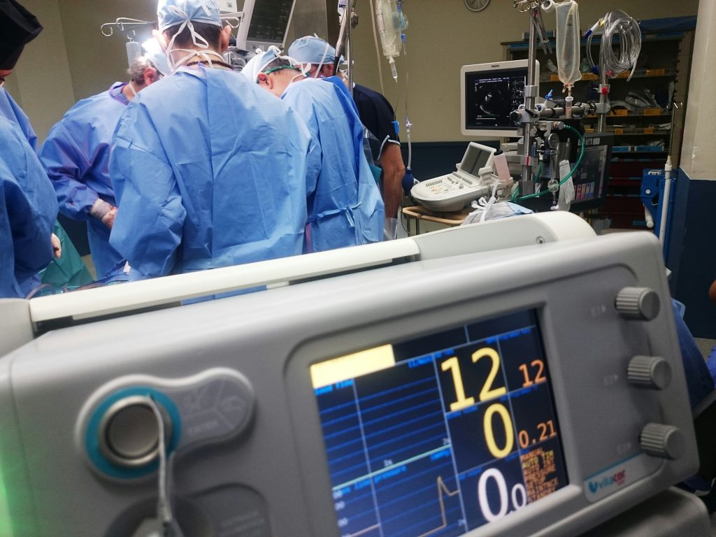 A team of surgeons performing an operation in an operating room. They are wearing blue surgical gowns, masks, and caps. In the foreground, there is a medical monitor displaying vital signs, and in the background, various medical equipment and IV lines are visible.